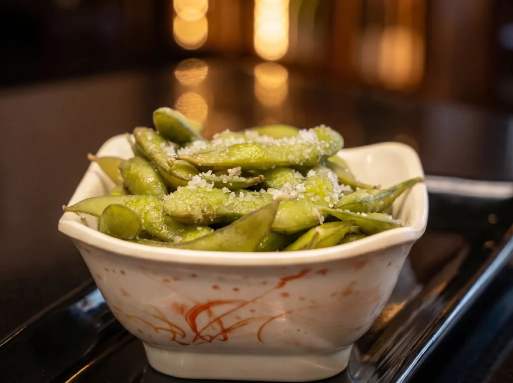 Steamed edamame pods sprinkled with coarse sea salt at Kingdom Sushi, served in a white square bowl with orange brushstroke accents