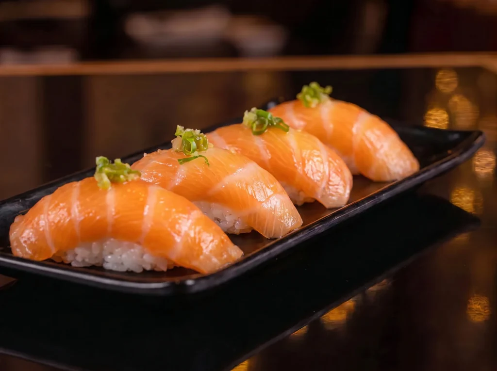 Salmon nigiri at Kingdom Sushi, four pieces of glossy salmon over rice topped with a dab of wasabi, on a black plate