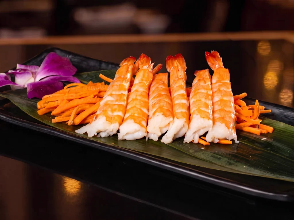 Shrimp Sashimi at Kingdom Sushi, whole prawns arranged over a green leaf with shredded carrots and a purple orchid on a black plate